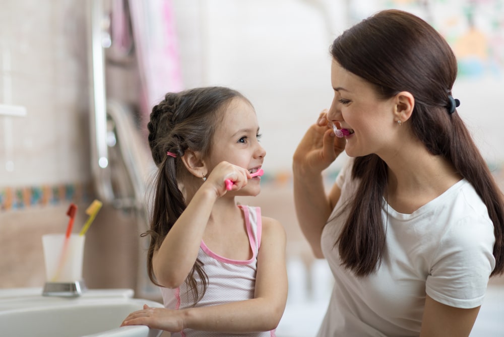 An adult woman and a young girl are standing in a bathroom, smiling and brushing their teeth together.