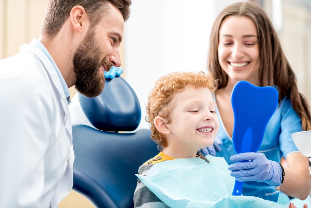 A young boy smiles at his reflection in a handheld mirror while sitting in a dental chair, with a dentist and dental assistant—experts in preventive dentistry—smiling beside him.