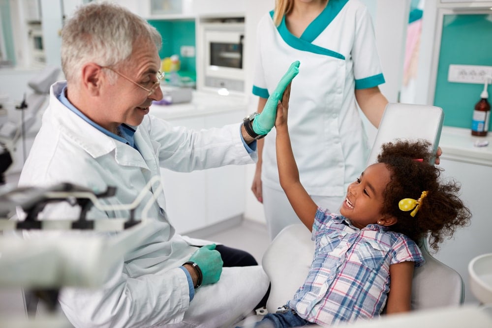 A dentist in a white coat gives a high-five to a smiling young girl sitting in a dental chair, highlighting the importance of Preventive Dentistry, while another dental staff member stands nearby.