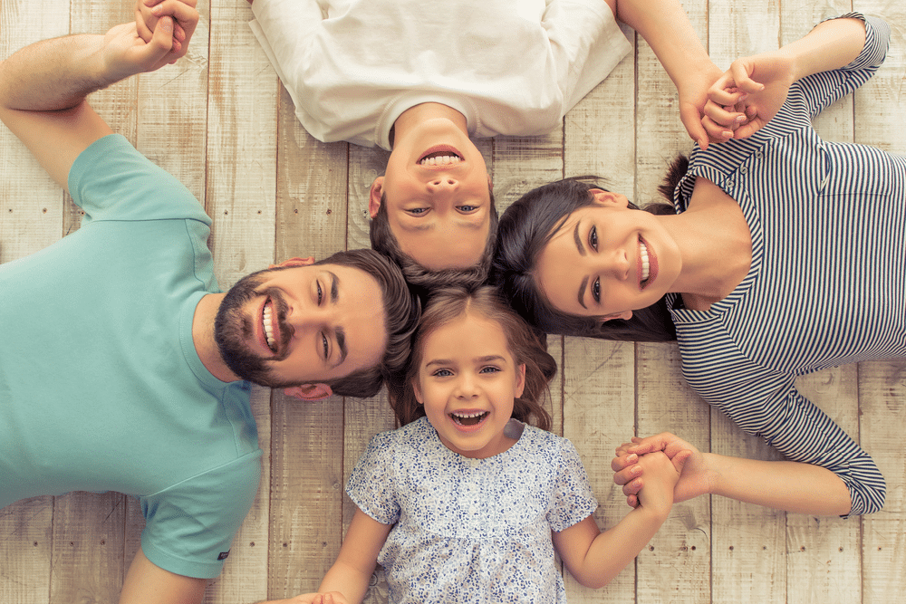 Four people, two adults and two children, lie on a wooden floor in a circle, holding hands and smiling up at the camera—perfect moments that start with healthy habits like pediatric dentistry.