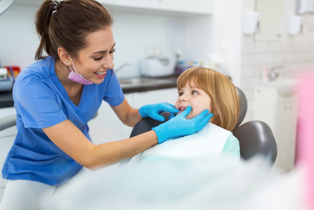 A dentist wearing blue scrubs examines a smiling young girl’s teeth in a dental office.