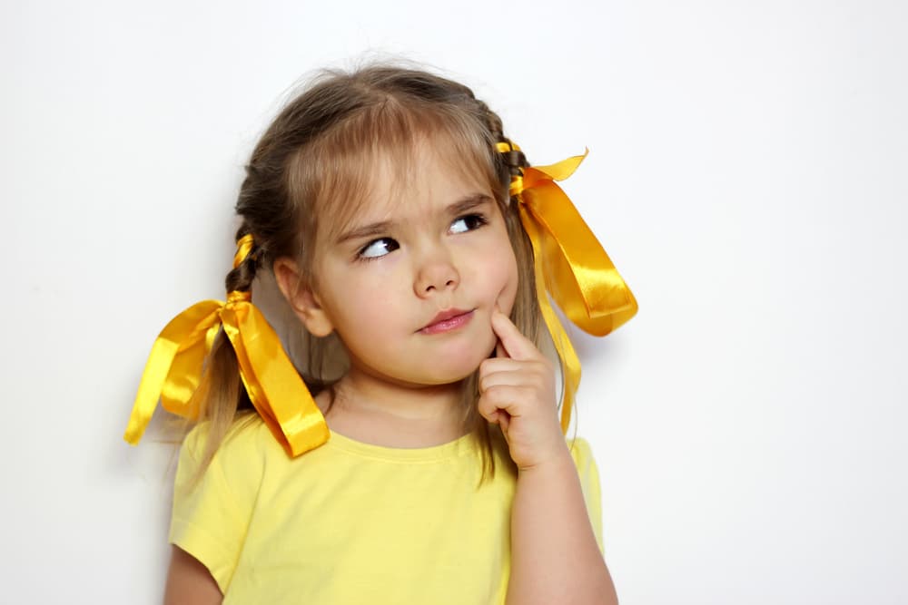 Young girl with yellow ribbons in her hair and a yellow shirt stands against a white background, touching her face and looking up thoughtfully.