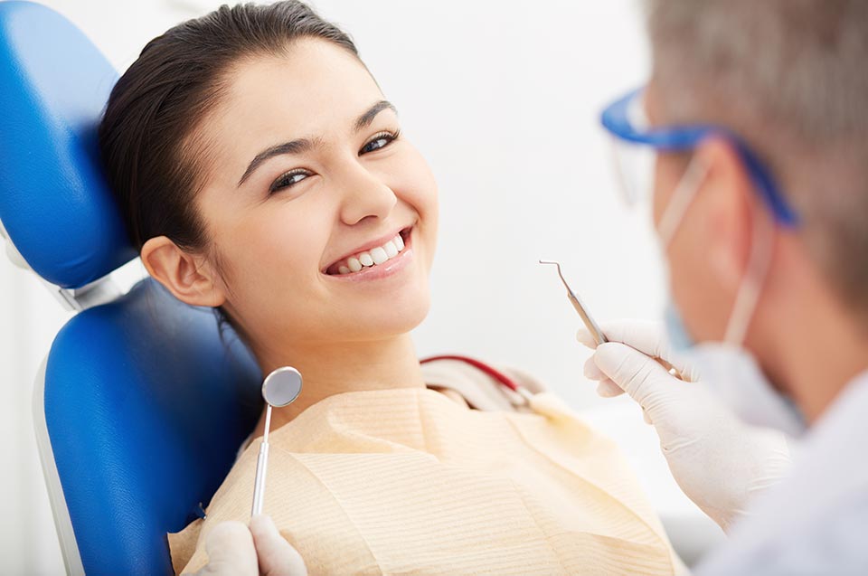 A woman sits in a dental chair smiling while a dentist, experienced in Sedation Dentistry, holds dental instruments and prepares for an examination.